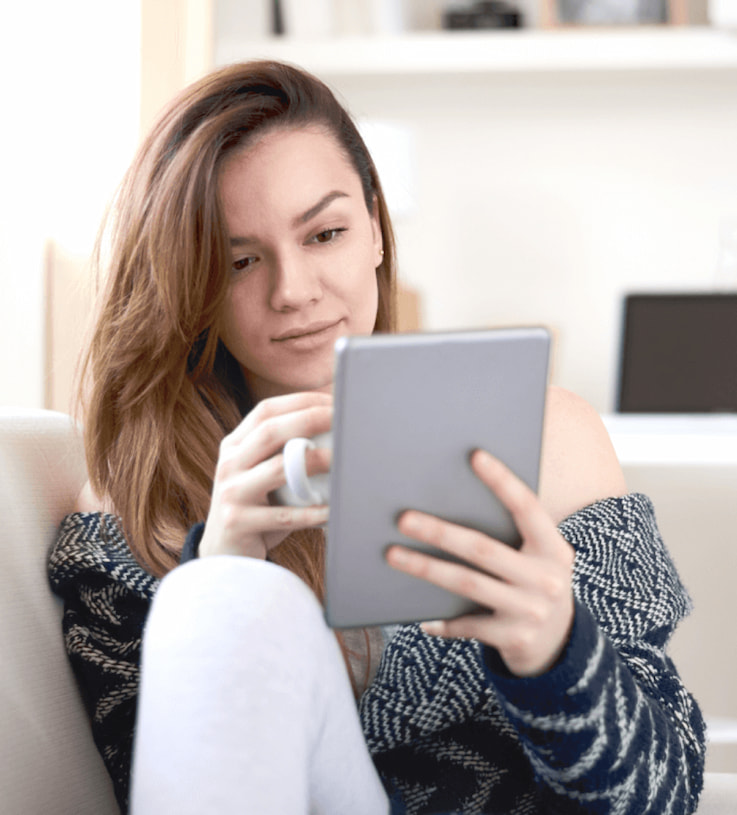 Woman sitting with a cup on one hand while holding and looking at a tablet on the other hand