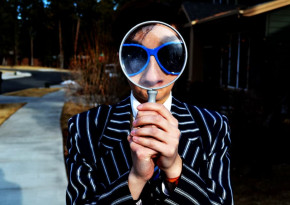 Man in blue suit with black stripes and blue sunglasses looking at the camera through a magnifying glass